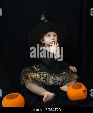 A little girl in a witch costume - a hat and a skirt with a spider web-sits on a black background and looks up in surprise. Next to it is a bucket-pum Stock Photo