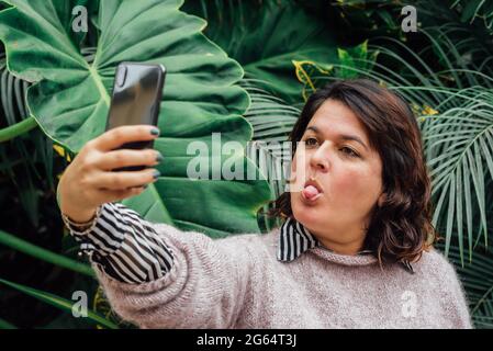 Woman sticks out her tongue in a video call and we see that she is wearing winter clothes and in a background with foliage Stock Photo