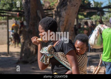 Mother carrying baby on her back, using a sarong cloth sling tied on her shoulders and eating raw cane sugar Stock Photo