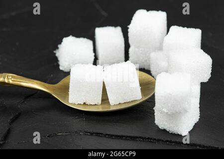 Several white sugar cubes were placed in a brass spoon on a black stone background. Stock Photo