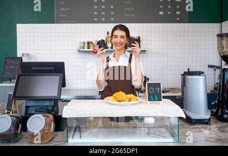 Young caucasian coffee shop staff  dressed in brown apron holding coffee drink in plastic cup with both hands. Morning atmosphere in a coffee shop. Stock Photo