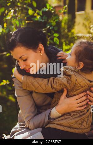 girl in a mustard-colored jacket hugs her mother sitting on a bench Stock Photo