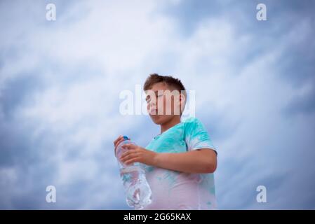 Boy with a bottle of clean water on the background of the sky. Thirst. Drink water. Stock Photo