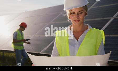 African-American engineer showing female technologist objects on paper diagram leaning on solar panel. Concept of green electricity. Stock Photo