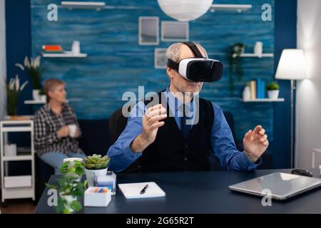 Senior man wearing virtual reality headset doing hand gesturing. Elderly man pensioner in living room sitting at desk with futuristic goggles for augmented cyberspace wife watching tv. Stock Photo