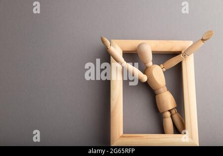 Wooden mannequin climbs out of a wooden frame with raised arms on gray pastel background. escape, break rules, freedom, welcome, greeting, joy concept Stock Photo