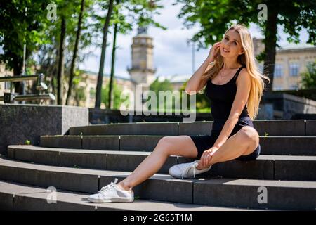 Young beautiful blonde sportive woman in a black tank top and in black tight sports shorts sits on the stairs Stock Photo