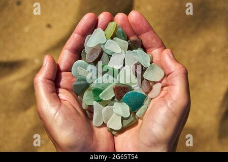 A person holds a collection of multi colored sea glass in their cupped hands with the sand in the background Stock Photo