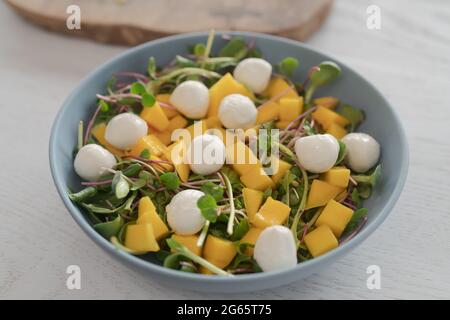 Salad with mango, mozzarella and mixed microgreens in blue bowl on white oak table, shallow focus Stock Photo