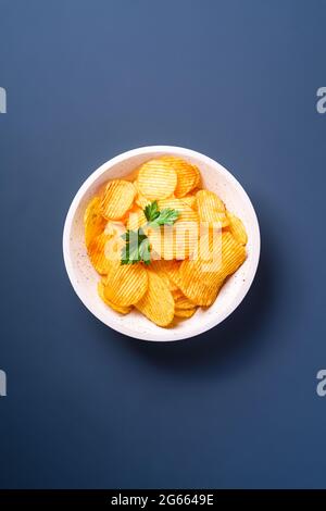 Fried corrugated golden potato chips with parsley leaf in wooden bowl on blue background, top view Stock Photo