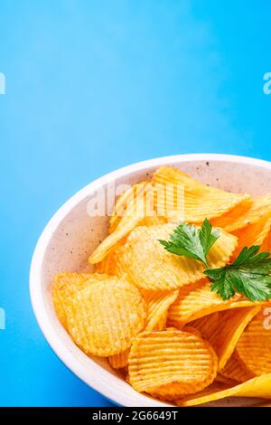 Fried corrugated golden potato chips with parsley leaf in wooden bowl on blue background, angle view macro Stock Photo
