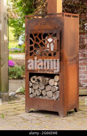 Rusty old wood stove with wood logs standing in a beautiful idyllic garden with fowers, trees and greenery on a sunny day creating a mindful scenery Stock Photo