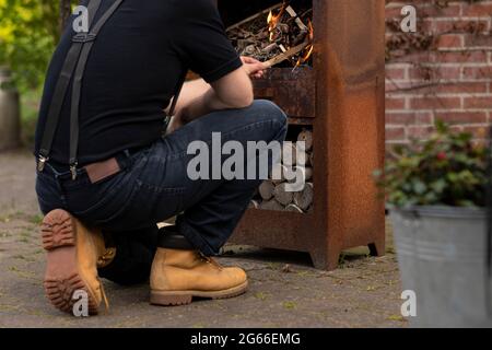 A man wearing suspenders making a fire with small wooden branches in a outdoor rusty old metal fire stove while relaxing and enjoying the simple life Stock Photo