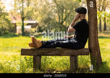 Farmer man wearing a cap and casual clothes sitting on a wooden bench calling with his mobile phone, working and doing business while looking over a g Stock Photo
