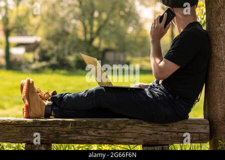Farmer man wearing a cap and casual clothes sitting on a wooden bench calling with his mobile phone, working on his laptop and doing business while lo Stock Photo
