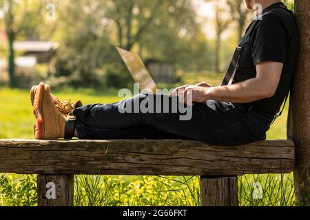 Digital nomad man wearing a cap and casual clothes sitting on a wooden bench calling with his mobile phone, working on his laptop and doing business w Stock Photo