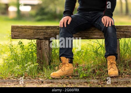 Man worker or farmer wearing casual clothes and shoes sitting, resting and relaxing, enjoying the nature outside on a rustic bench next to a green gra Stock Photo
