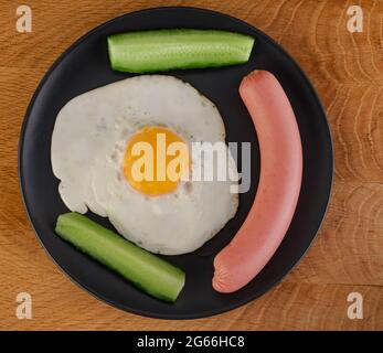 Scrambled eggs, sausage, cucumbers in a black plate on a wooden board Stock Photo
