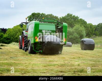 Hay or silage making (farmer & farm tractor at work in rural field, collecting grass, wrapped round bale dropping from baler) - Yorkshire England, UK. Stock Photo