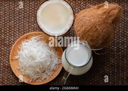 High angle view of coconut products, such as, whole brown coconut, coconut shredded and coconut milk. On dark brown bamboo weave textured background. Stock Photo