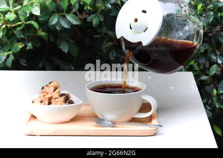 Pour coffee from pot into cup. Coffee cup served on wood tray alongside with cookies. Table set in garden atmosphere with green leaves background. Stock Photo