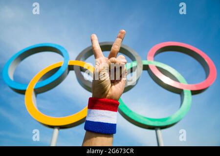 RIO DE JANEIRO - CIRCA MAY, 2016: A hand wearing red white and blue wristband makes a victory sign in front of Olympic Rings standing under bright blu Stock Photo