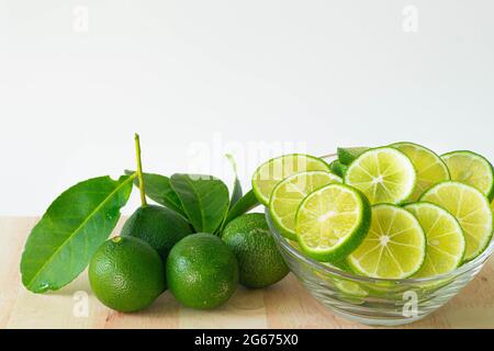 Green limes with leaves and sliced lime in glass bowl on wood, white background. Copy space. Stock Photo
