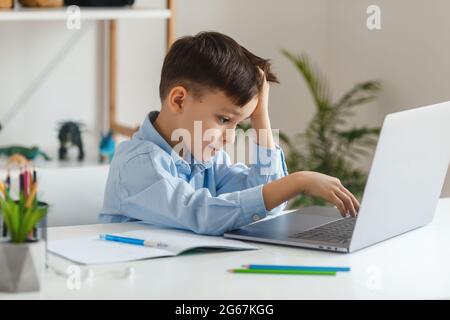 Clever kid tired to learn difficult homework using laptop and internet. Elementary school e-learning via video call. Boy studying online at home Stock Photo