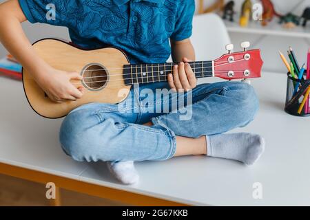 Close-up kid playing soprano ukulele sitting on desk. Preschool boy learning guitar at leisure. Concept of early childhood education and music hobby Stock Photo