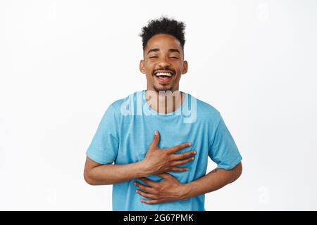 Image of candid happy african american guy laughing, holding hands on chest and belly, chuckle over smth funny, watching comedy, standing over white Stock Photo