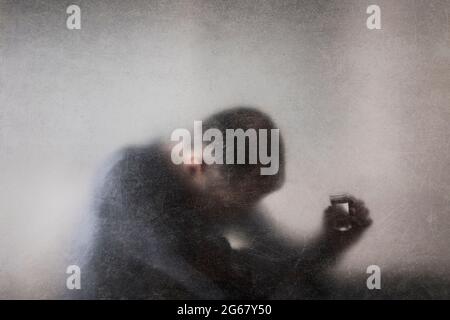 Man with a glass of vodka behind a dusty scratched glass. Alcoholism concept. Stock Photo