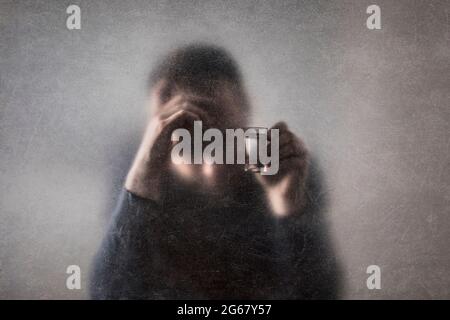 Man with a glass of vodka behind a dusty scratched glass. Alcoholism concept. Stock Photo