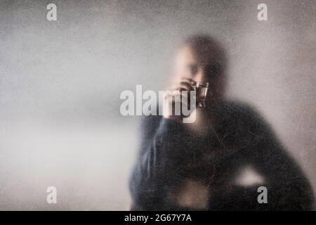 Man with a glass of vodka behind a dusty scratched glass. Alcoholism concept. Stock Photo