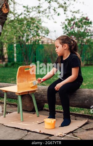 a girl sits on a log in the garden and paints outdated broken furniture in a new color, a family business, a restoration workshop Stock Photo