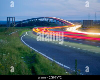 Elmley, Kent, UK. 3rd July 2021. Today marks the 15th anniversary of the Sheppey Crossing in Kent opening. The bridge opened 3 July 2006 as part of a £100m improvement plan for the A249 between the M2 and Sheerness Port (a key importer of cars). Kent Online reports Highways England as saying an average of 34,060 vehicles have used the crossing daily in the past 12 months. Pic: in-camera composite long exposure showing car light trails, the older Kingsferry Bridge can be seen on the left of the picture. Credit: James Bell/Alamy Live News Stock Photo