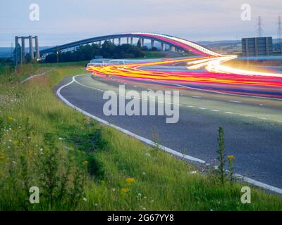 Elmley, Kent, UK. 3rd July 2021. Today marks the 15th anniversary of the Sheppey Crossing in Kent opening. The bridge opened 3 July 2006 as part of a £100m improvement plan for the A249 between the M2 and Sheerness Port (a key importer of cars). Kent Online reports Highways England as saying an average of 34,060 vehicles have used the crossing daily in the past 12 months. Pic: in-camera composite long exposure showing car light trails, the older Kingsferry Bridge can be seen on the left of the picture. Credit: James Bell/Alamy Live News Stock Photo