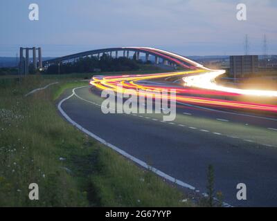 Elmley, Kent, UK. 3rd July 2021. Today marks the 15th anniversary of the Sheppey Crossing in Kent opening. The bridge opened 3 July 2006 as part of a £100m improvement plan for the A249 between the M2 and Sheerness Port (a key importer of cars). Kent Online reports Highways England as saying an average of 34,060 vehicles have used the crossing daily in the past 12 months. Pic: in-camera composite long exposure showing car light trails, the older Kingsferry Bridge can be seen on the left of the picture. Credit: James Bell/Alamy Live News Stock Photo