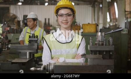 Portrait Asian engineer woman worker wear safety goggles control lathe machine industrial manufacturing factory crossed arm smiling happy looking came Stock Photo