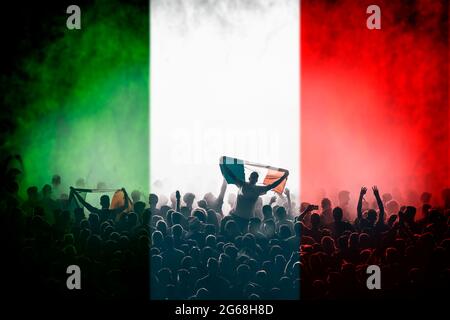 Soccer fans supporting Italy - crowd celebrating in stadium with raised hands against italian flag Stock Photo