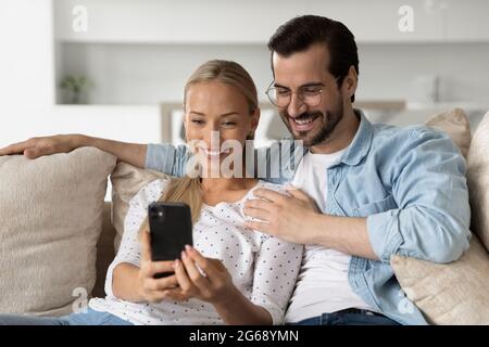 Smiling young couple hugging using smartphone sitting on couch together Stock Photo