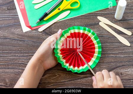 In the hands of a child Paper Fan watermelon on a wooden table. Childrens art project, handmade, crafts for children. Stock Photo