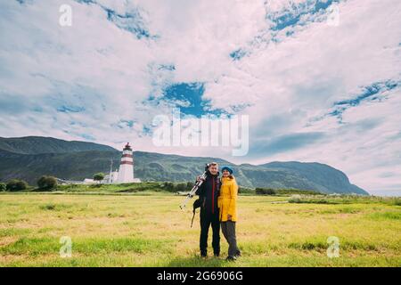 Alnesgard, Godoya, Norway. Man And Woman Young Adult Caucasian Tourists Travelers Couple Posing Against Old Alnes Lighthouse In Summer Day In Godoy Stock Photo
