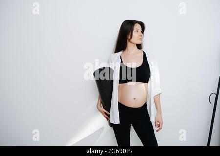 Portait of a pregnant woman holding yoga mat in hand. She's wearing black top and pants. Looking to the side.