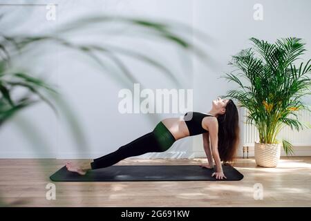Side view on a pregnant woman practicing yoga alone, doing reverse plank in a big studio. She's wearing black top and pants Stock Photo