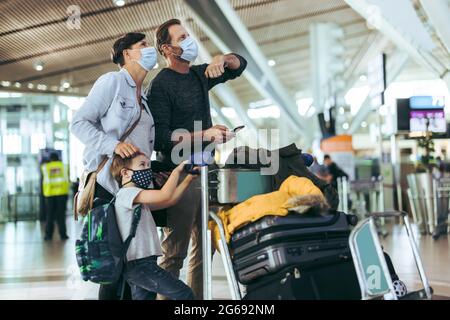 Family in face masks looking at their flight times on board at airport. Tourist man with wife and kid waiting at airport with luggage on trolley durin Stock Photo