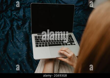 Top view of a woman working on a laptop with a blank black screen while sitting on a cozy bed at hom Stock Photo