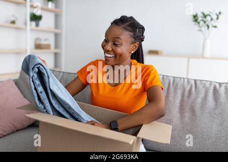 Unpacking clothes. Portrait of smiling black lady opening cardboard box, taking out new jeans, sitting on sofa at home Stock Photo