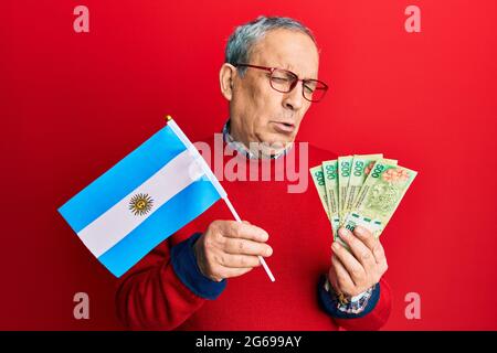 Handsome senior man with grey hair holding argentine flag and pesos banknotes depressed and worry for distress, crying angry and afraid. sad expressio Stock Photo