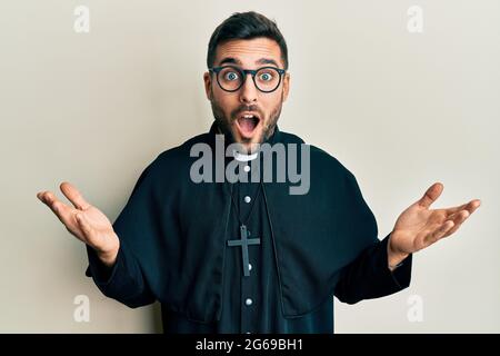 Young hispanic priest man standing over white background afraid and shocked with surprise and amazed expression, fear and excited face. Stock Photo