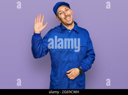 Bald man with beard wearing builder jumpsuit uniform waiving saying hello happy and smiling, friendly welcome gesture Stock Photo